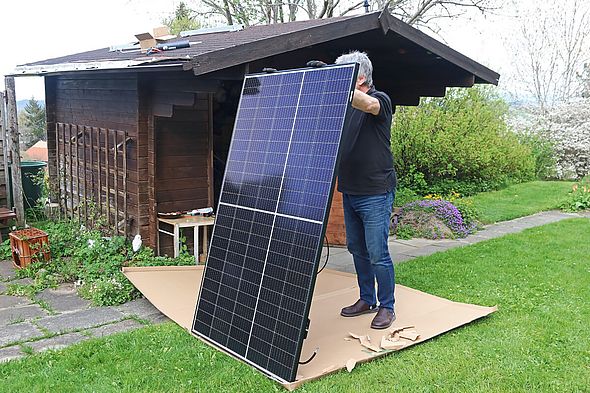 Mann steht vor einem Gartenhaus und hält ein Solarmodul auf dem Rasen