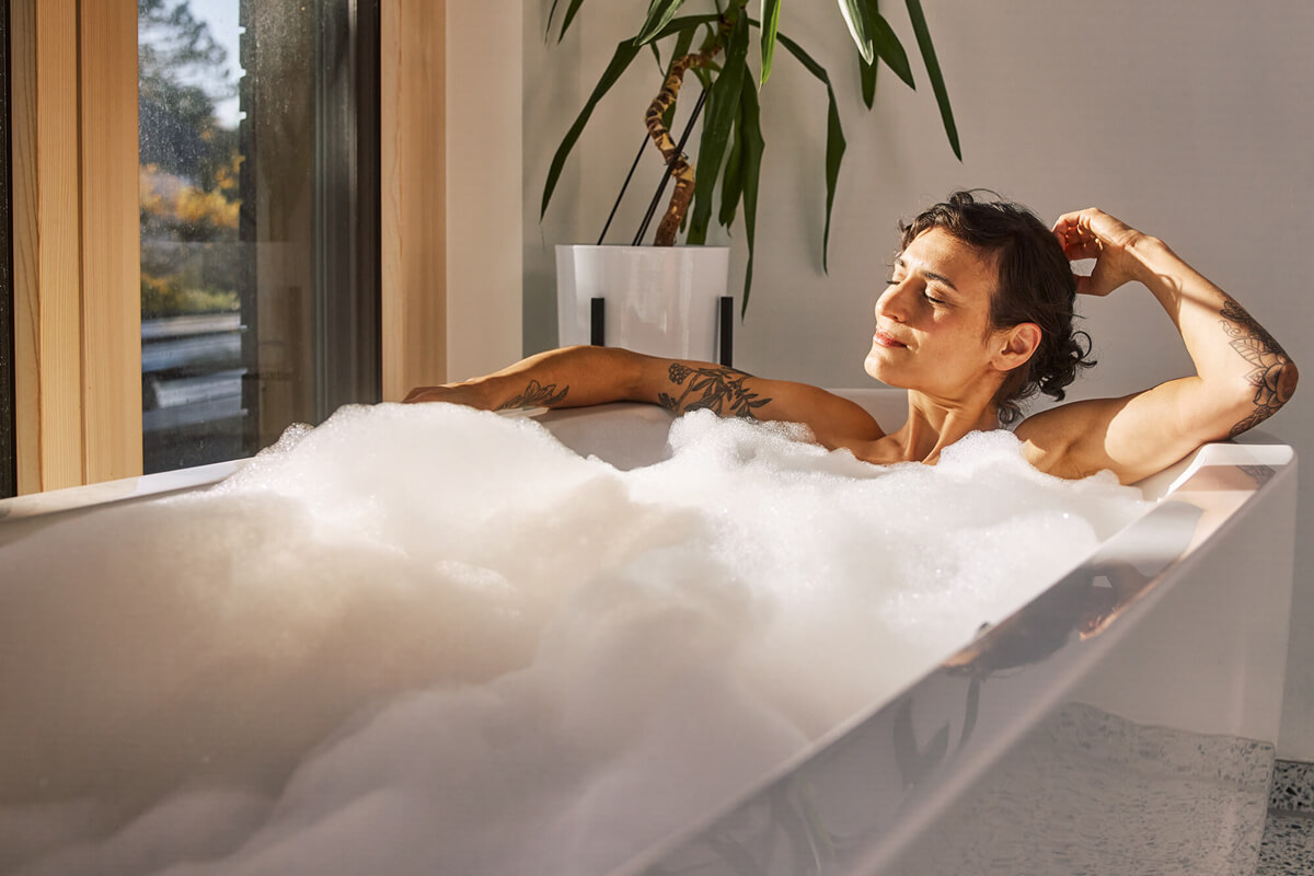 Entpannt aussehende Frau mit Tattoos auf den Armen in einer Badewanne voller Schaum, im Hintergrund ein Fenster und eine Zimmerpflanze