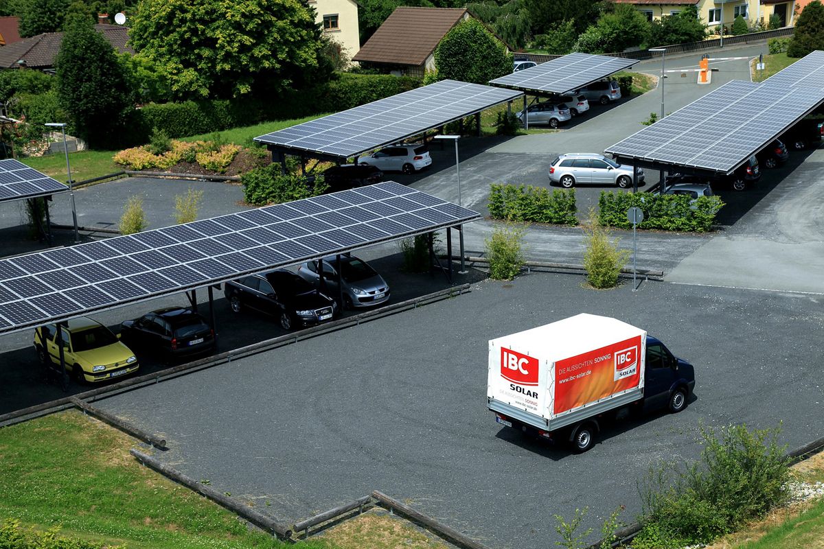 Schotterparkplatz mit Carports, die Solarmodule auf dem Dach haben; im Vordergrund ein kleiner Laster, der mit IBC SOLAR Werbung bedruckt ist