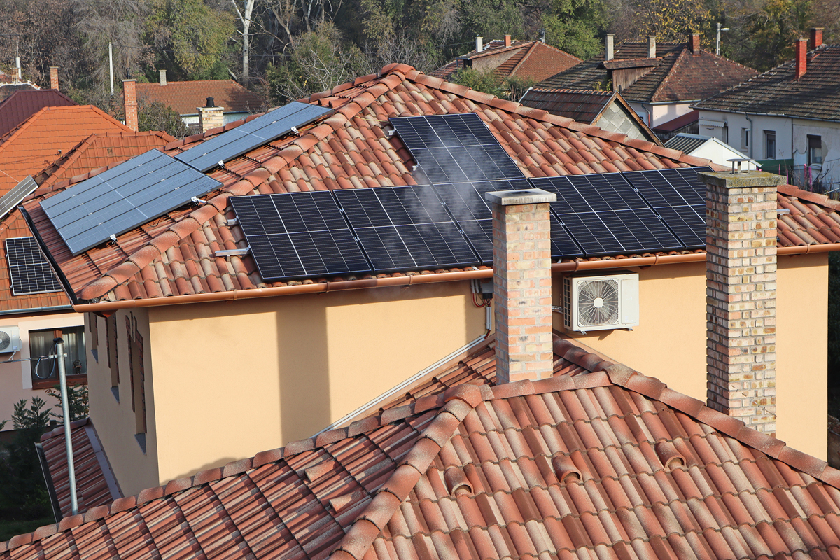 Mehrere Hausdächer aus der Vogelperspektive, auf zwei Dächern befinden sich Solarmodule, ein Schornstein im Vordergrund raucht und an der Wand eines Hauses ist eine Klimaanlage montiert