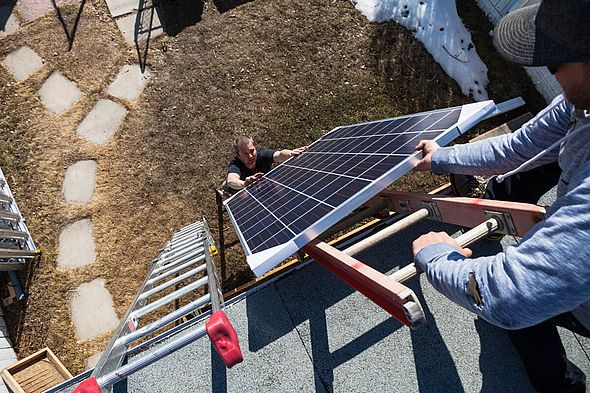 Zwei Menschen davon einer auf dem Dach der ander auf dem Boden, zwischen ihnen eine Leiter und ein Solarmodul, das von unten nach oben gereicht wird