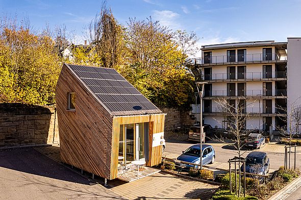Tiny House aus Holz mit Solarmodulen auf dem Dach auf einem Parkplatz; im Hintergrund geparkte Autos, ein Haus und herbstliche Bäume