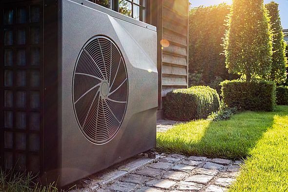 Grüner Garten mit Hecken und Bäumen in den die Sonne fällt mit grauer Wärmepumpe im Vordergrund