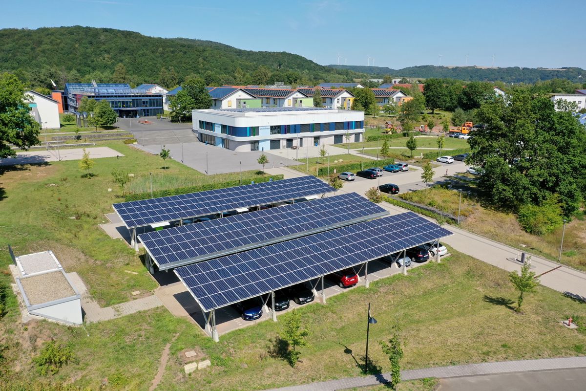 Solar-Carports mit Photovoltaikanlagen auf Parkplätzen vor einem Bürogebäude; im Hintergrund Wohnhäuser mit Solardächern.